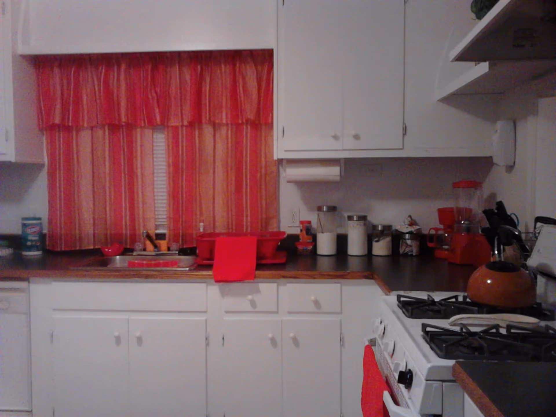 A kitchen with white cabinets and dark countertops. There is a window with red and orange striped curtains above the sink. On the counter, there are various kitchen items including a red dish rack, containers with white ingredients, a red blender, and a red kettle on a white gas stove. A red towel hangs on the oven door.