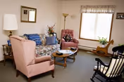 A cozy living room with two pink upholstered armchairs, a floral patterned sofa with a blue throw blanket, a wooden coffee table, a rocking chair, a floor lamp, and a window with a floral valance letting in natural light. There are decorative items including flowers on the coffee table and a guitar near the window.