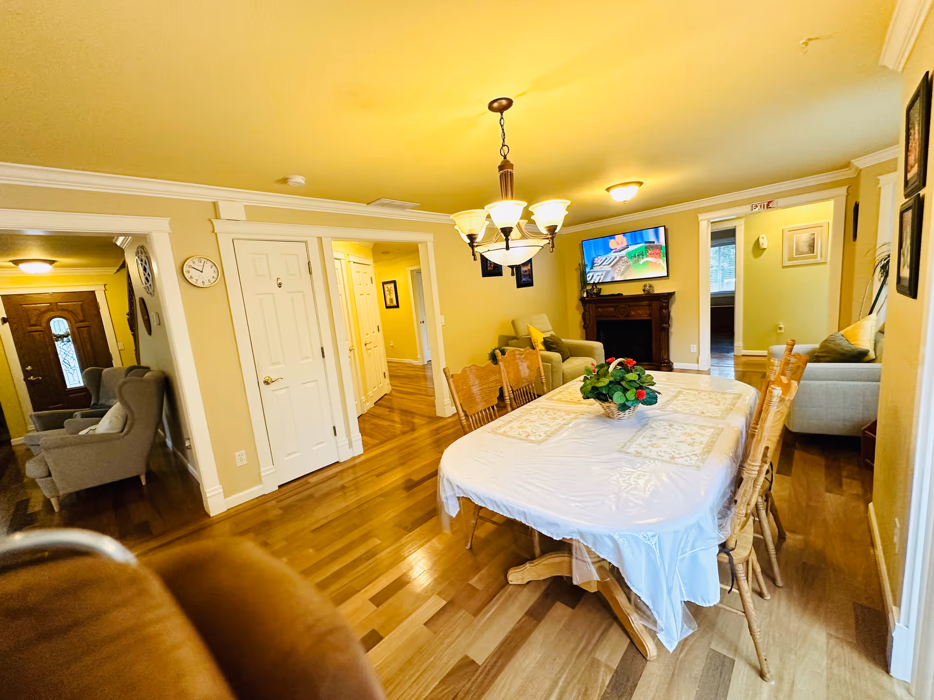 A warm, inviting dining room with a wooden table covered by a white tablecloth and six wooden chairs. A floral centerpiece decorates the table. The room has hardwood floors and yellow walls. A chandelier with multiple lights hangs above the table. In the background, there is a living area with armchairs, a wall-mounted TV above a fireplace, and an exit sign above a doorway.