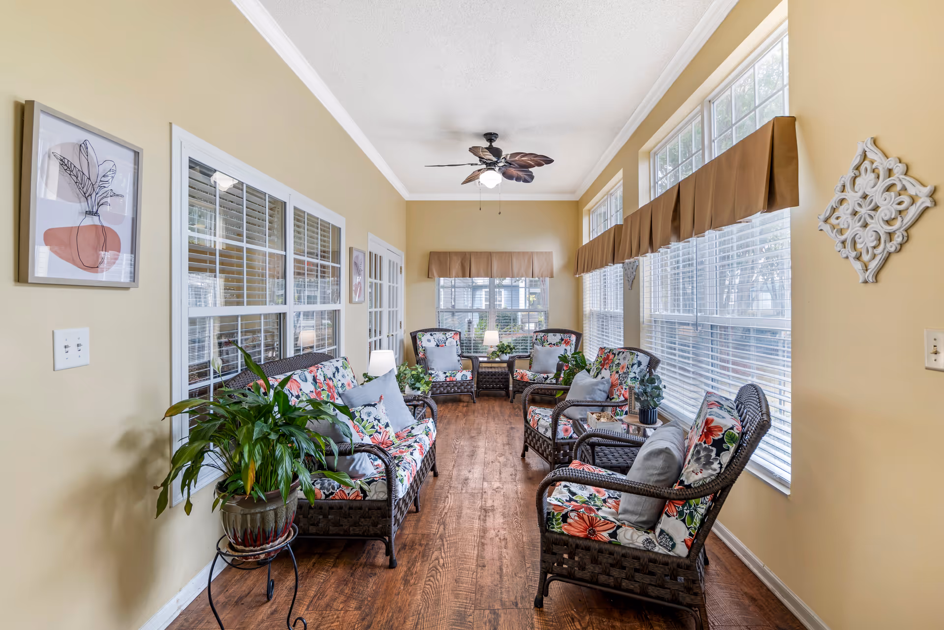 A bright sunroom with large windows on two sides, featuring wicker furniture with floral cushions, several potted plants, a ceiling fan with leaf-shaped blades, and light yellow walls decorated with framed artwork and a decorative wall piece.
