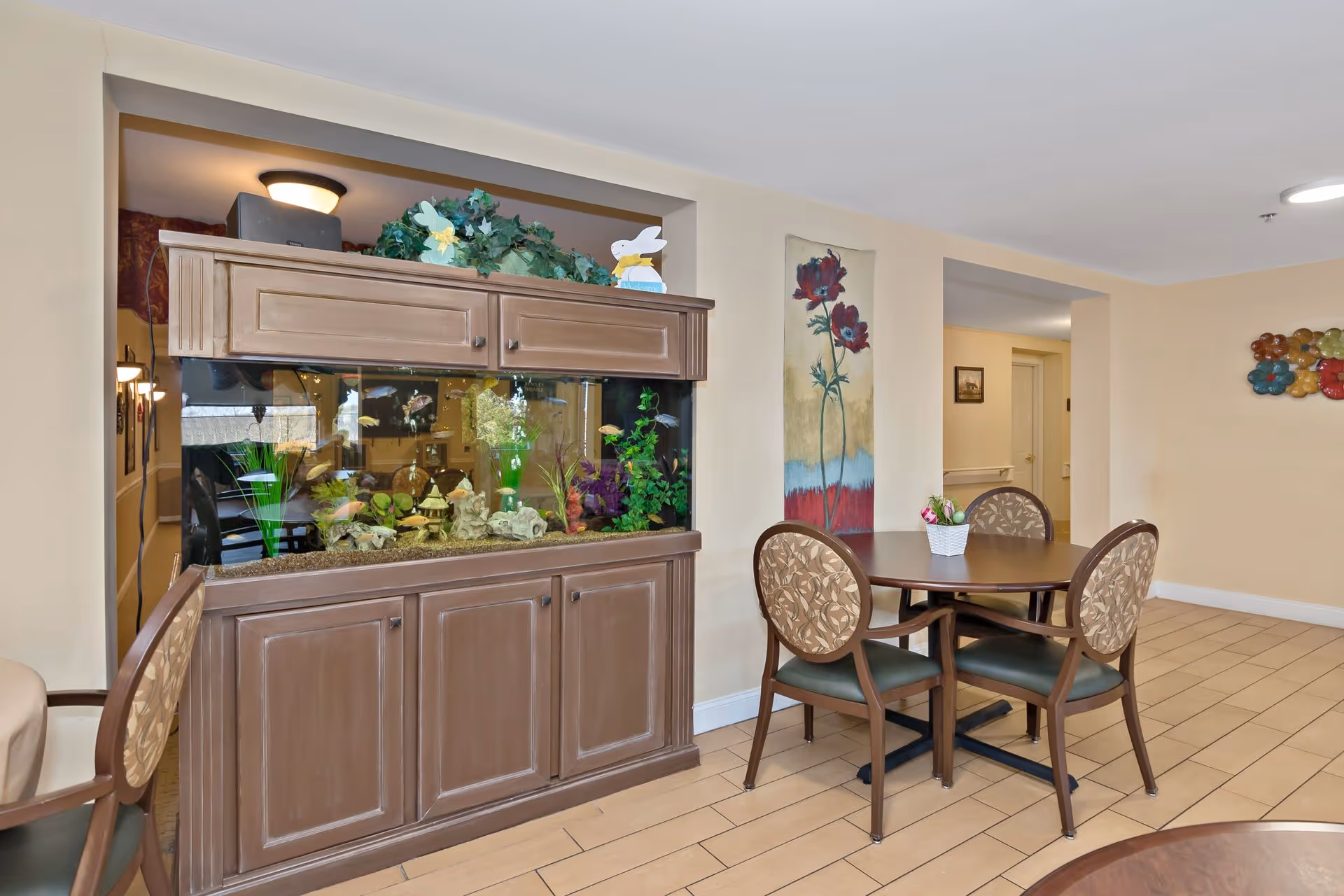 A cozy interior area of Bentley Assisted Living at Branchville featuring a large built-in fish tank with colorful fish and decorative plants. In front of the fish tank is a round wooden table with four cushioned chairs, and a small flower arrangement on the table. The walls are light beige with floral artwork and a decorative wall piece. The floor is tiled with light brown tiles.