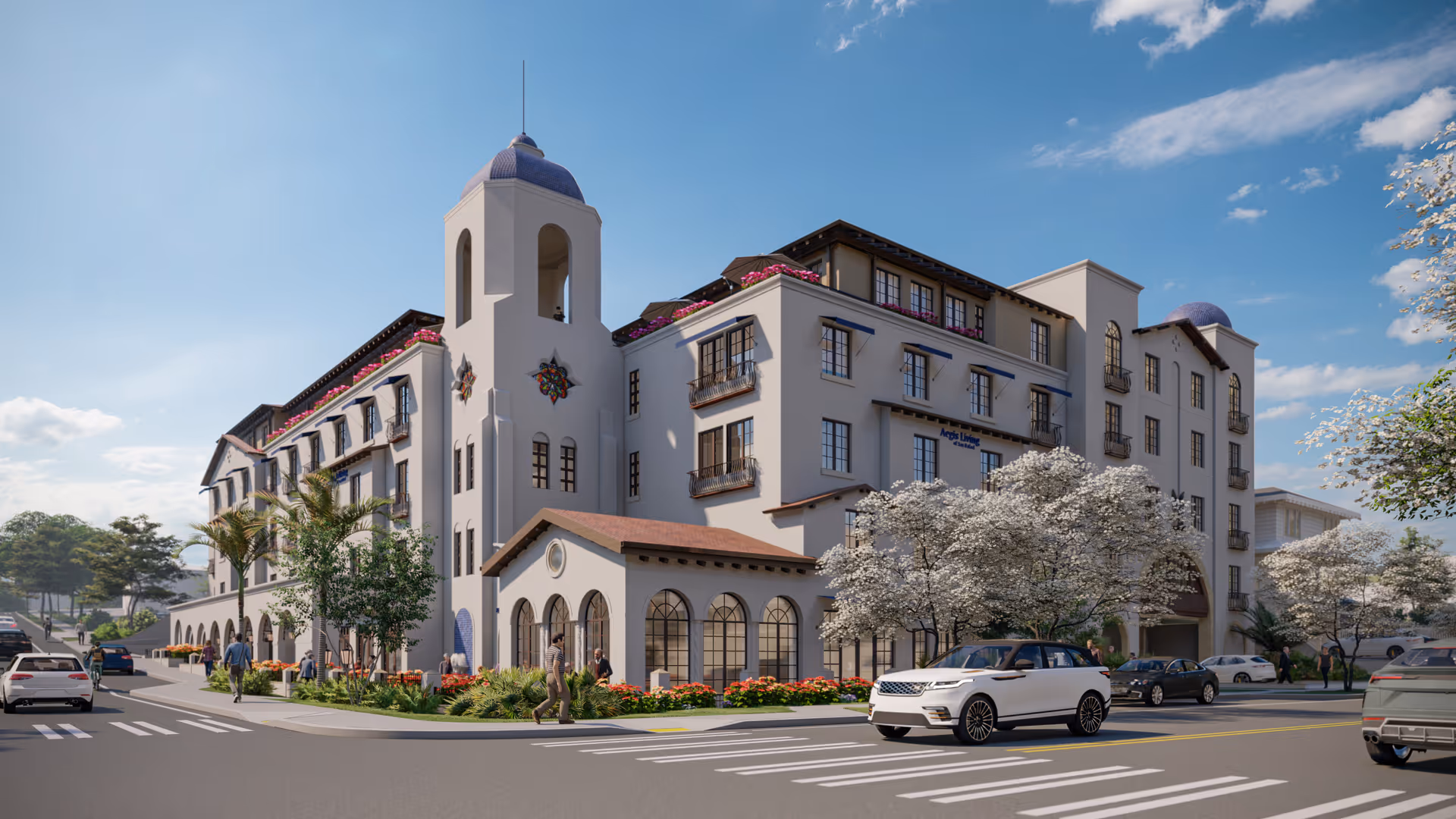 Exterior view of a large, multi-story senior living facility named Aegis Living San Rafael with a Mediterranean architectural style, featuring arched windows, balconies with flower boxes, and a tower with a blue dome. Several people are walking on the sidewalks and cars are driving on the street in front of the building under a clear blue sky.