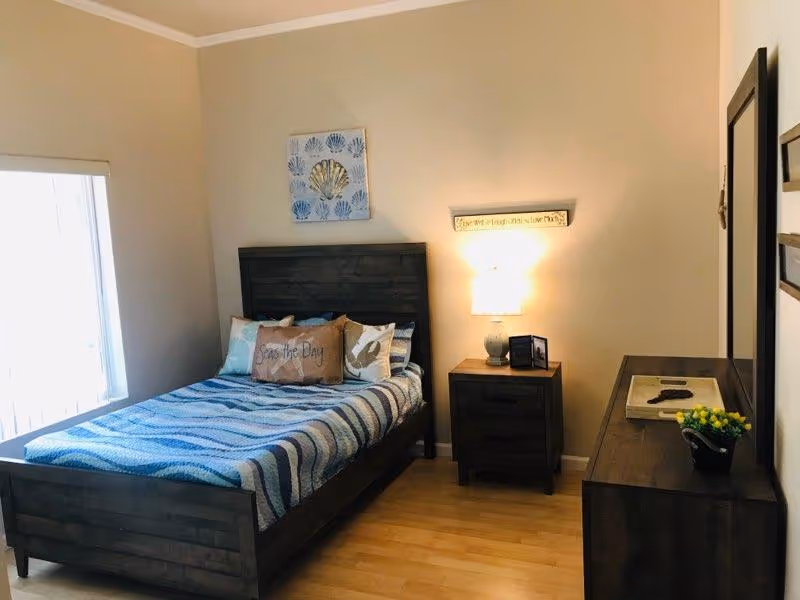 A cozy bedroom with a wooden bed frame and a blue and white striped blanket. The bed has several decorative pillows, including one that says 'Seas the Day.' There is a wooden nightstand with a lamp and a small picture frame next to the bed. A wooden dresser with a mirror and a small potted plant is on the right side of the room. The walls are light beige, and there is a window with blinds on the left side.