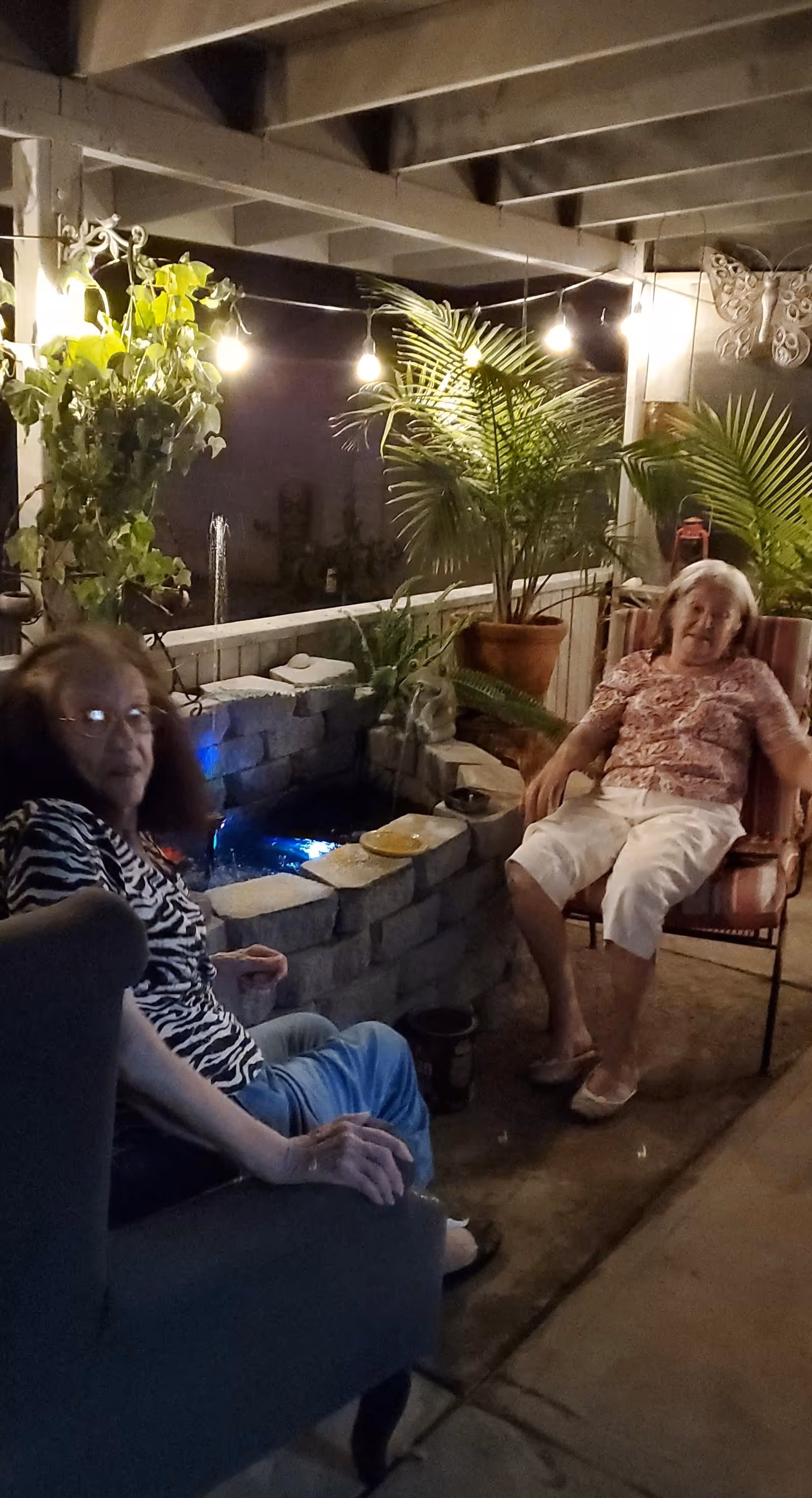 Two elderly women sitting and relaxing on a covered patio at night. The patio is decorated with potted plants, string lights, and a small water feature made of stacked stones. One woman is seated on a cushioned chair, and the other is seated on a different chair. The atmosphere is cozy and calm.