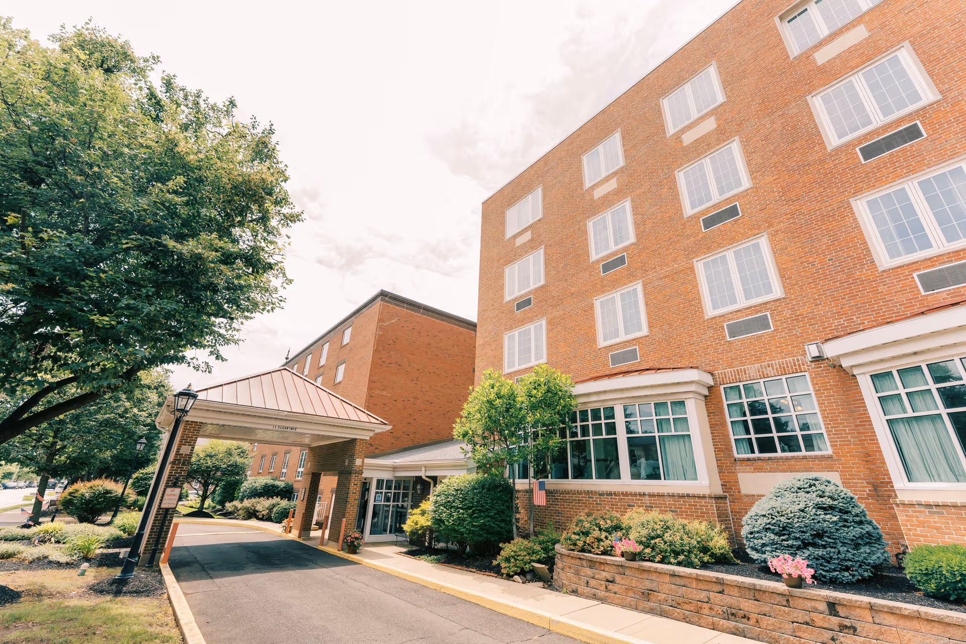 Exterior view of a multi-story brick building with large windows and a covered entrance driveway. The building is surrounded by well-maintained landscaping including bushes, flowers, and trees under a partly cloudy sky.