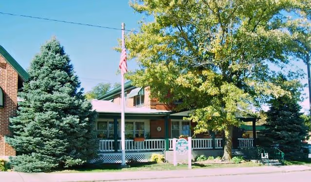 Exterior view of a brick senior living facility with a porch, white railing, an American flag on a flagpole, and large evergreen and deciduous trees in front.