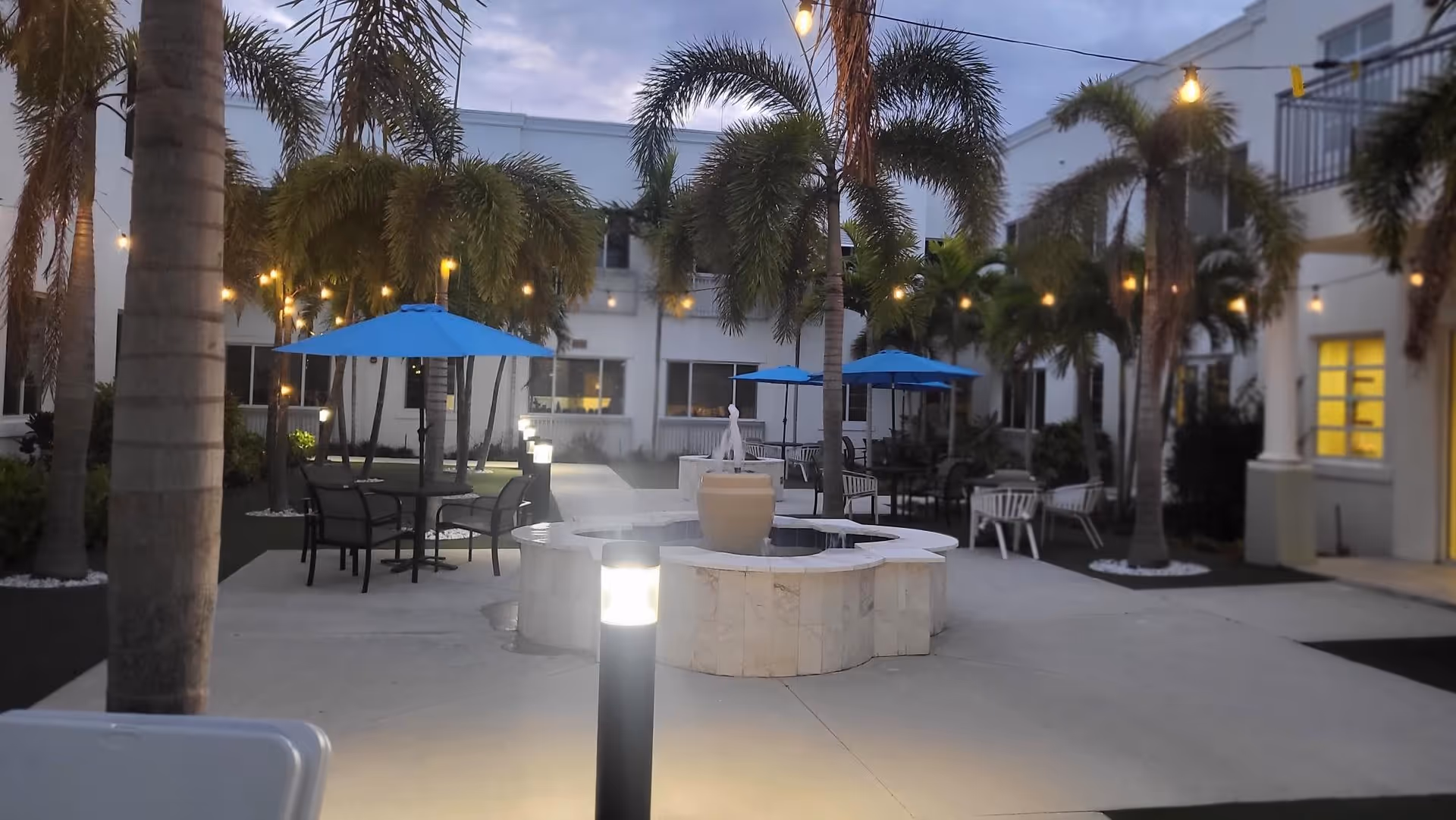 Outdoor courtyard area at dusk with palm trees, string lights, blue umbrellas over tables with chairs, and a central water fountain surrounded by a circular stone bench.
