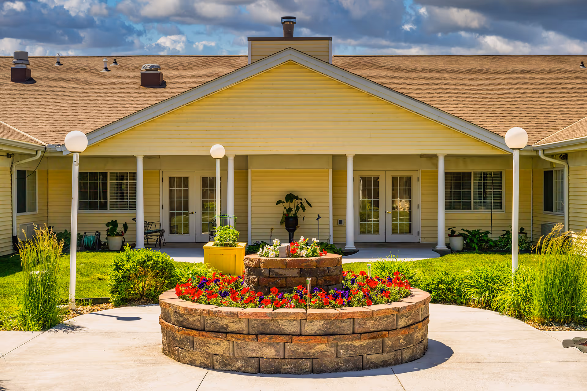 Outdoor courtyard area of a senior living facility with a circular stone flower bed filled with red and purple flowers in the center. The building has yellow siding, white columns, and multiple glass doors and windows. There are green plants and grass surrounding the flower bed, and four white globe lamp posts around the courtyard.