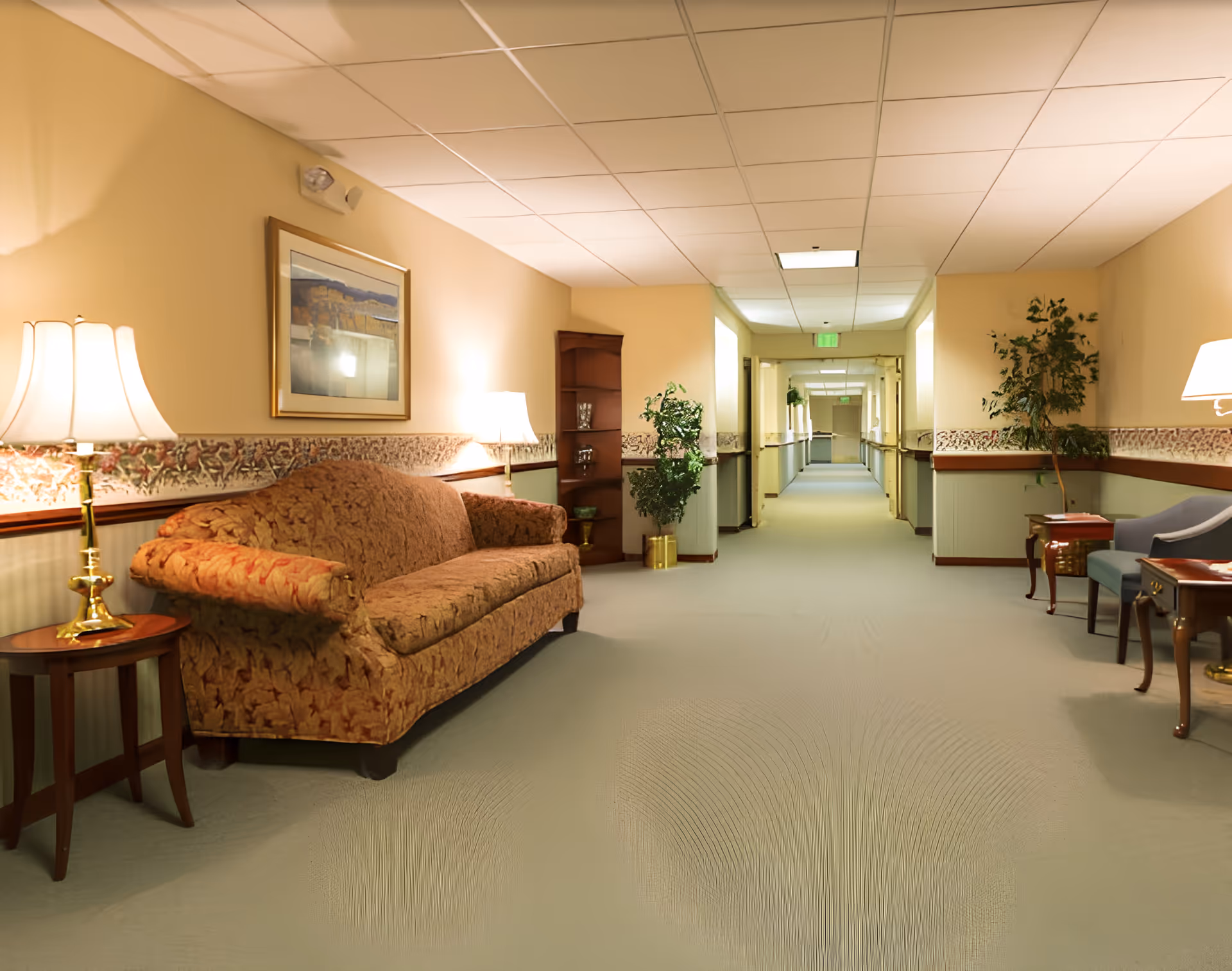 A well-lit hallway in a senior living facility with beige walls and carpeted floor. The hallway features a patterned wallpaper border, a brown patterned sofa with a side table and lamp on the left, and a blue armchair with a side table and lamp on the right. There are potted plants and framed artwork on the walls, with ceiling lights illuminating the corridor extending into the distance.