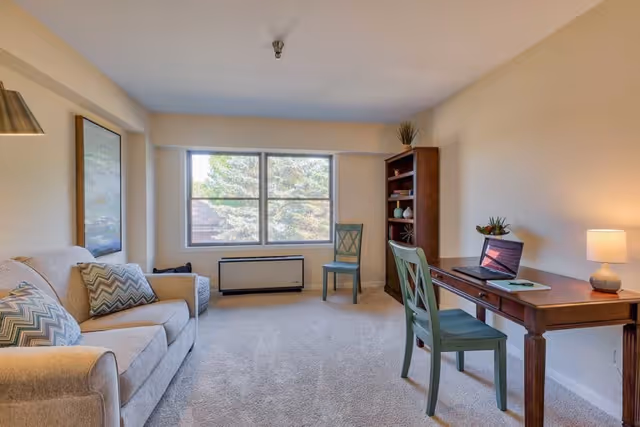 A bright living room with a beige sofa adorned with patterned pillows on the left, a wooden desk with a laptop and a small lamp on the right, two green chairs, a wooden bookshelf with decorative items, and a large window showing trees outside.