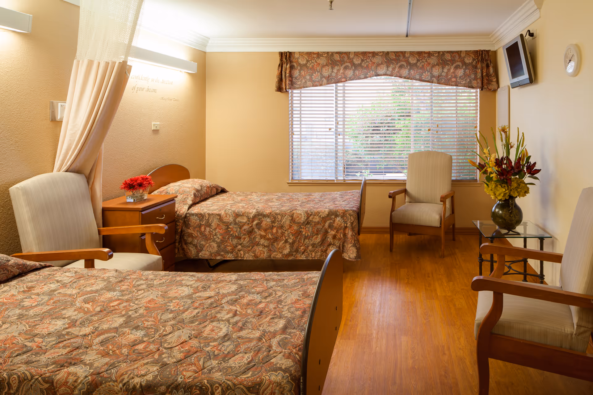 A cozy bedroom in a senior living facility with two single beds covered in patterned bedspreads. There are two armchairs, a wooden nightstand with a vase of red flowers, a glass side table with a vase of yellow and red flowers, a wall-mounted TV, and a large window with blinds and a valance. The room has warm beige walls and wooden flooring.