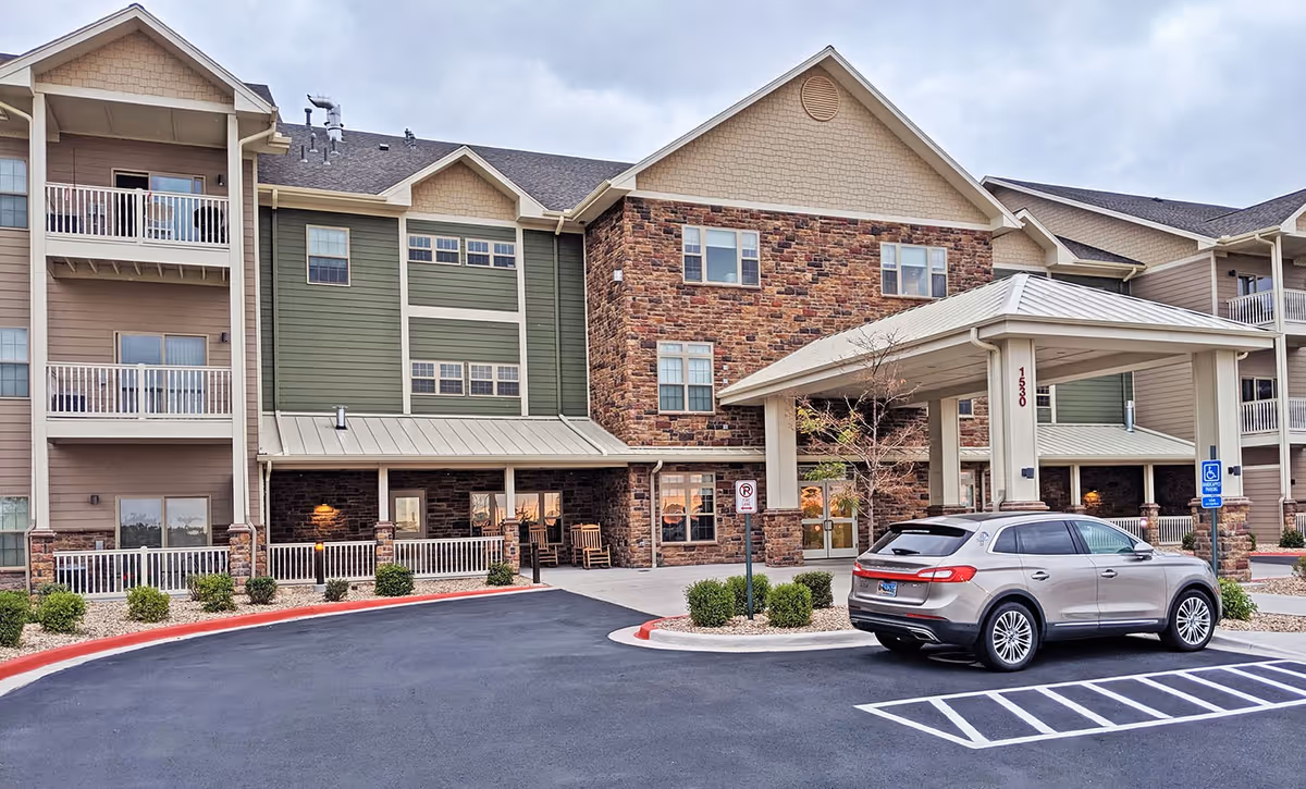 Exterior view of a multi-story senior living facility with a covered entrance, balconies, and a parked silver SUV in front. The building features a mix of stone and siding with green and beige colors, and there are small shrubs and landscaping around the entrance area.