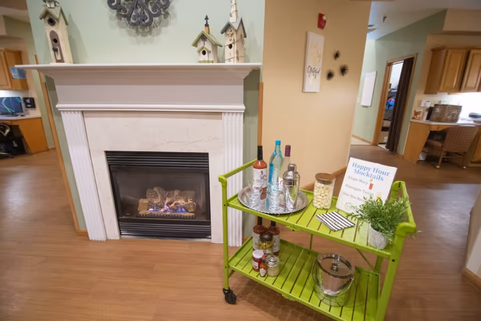 A cozy interior common area with a lit fireplace and a bright green cart holding mocktail bottles and supplies.