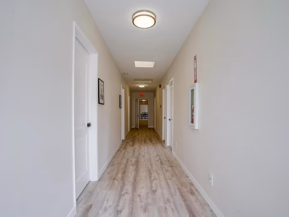 A long, narrow hallway with light wood flooring and beige walls. There are several white doors on both sides of the hallway, a fire extinguisher cabinet mounted on the right wall, and ceiling lights providing illumination. At the end of the hallway, a room with a table and chairs is visible.