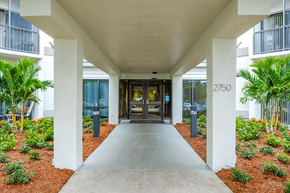 Covered entrance walkway leading to glass double doors of a building with the number 2750 on a white pillar, surrounded by landscaped greenery and palm plants.