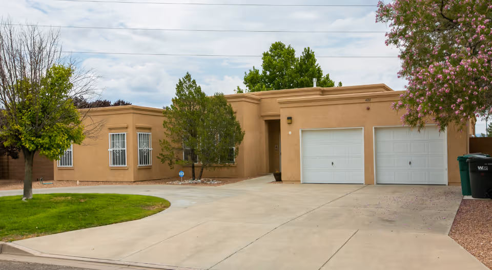 Single-story tan stucco building with two white garage doors, driveway, and surrounding trees.
