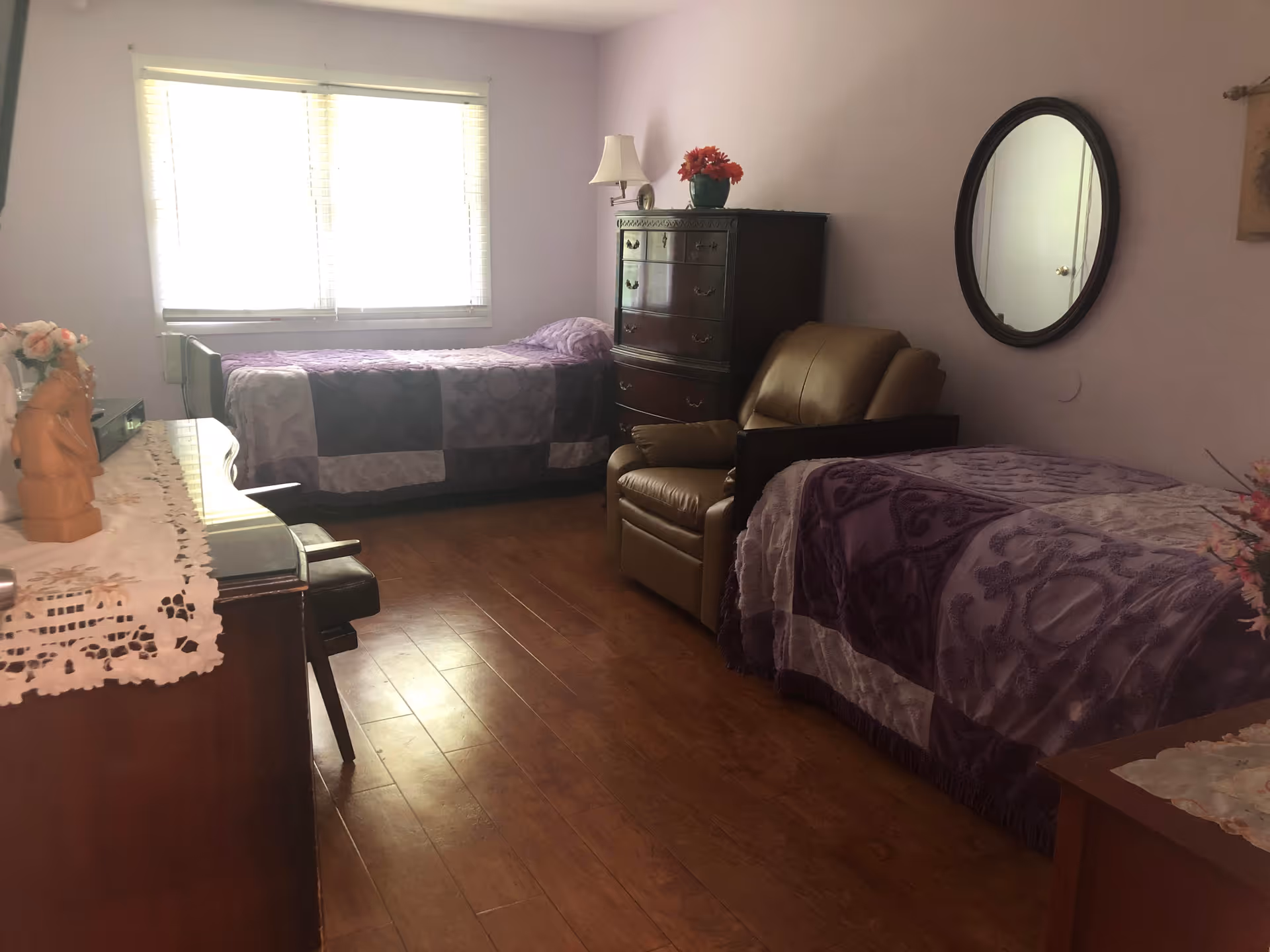 A bedroom with two single beds covered in purple patterned bedspreads, a brown leather armchair between the beds, a wooden dresser with a lamp and a flower pot, a round mirror on the wall, and a window with blinds letting in natural light.