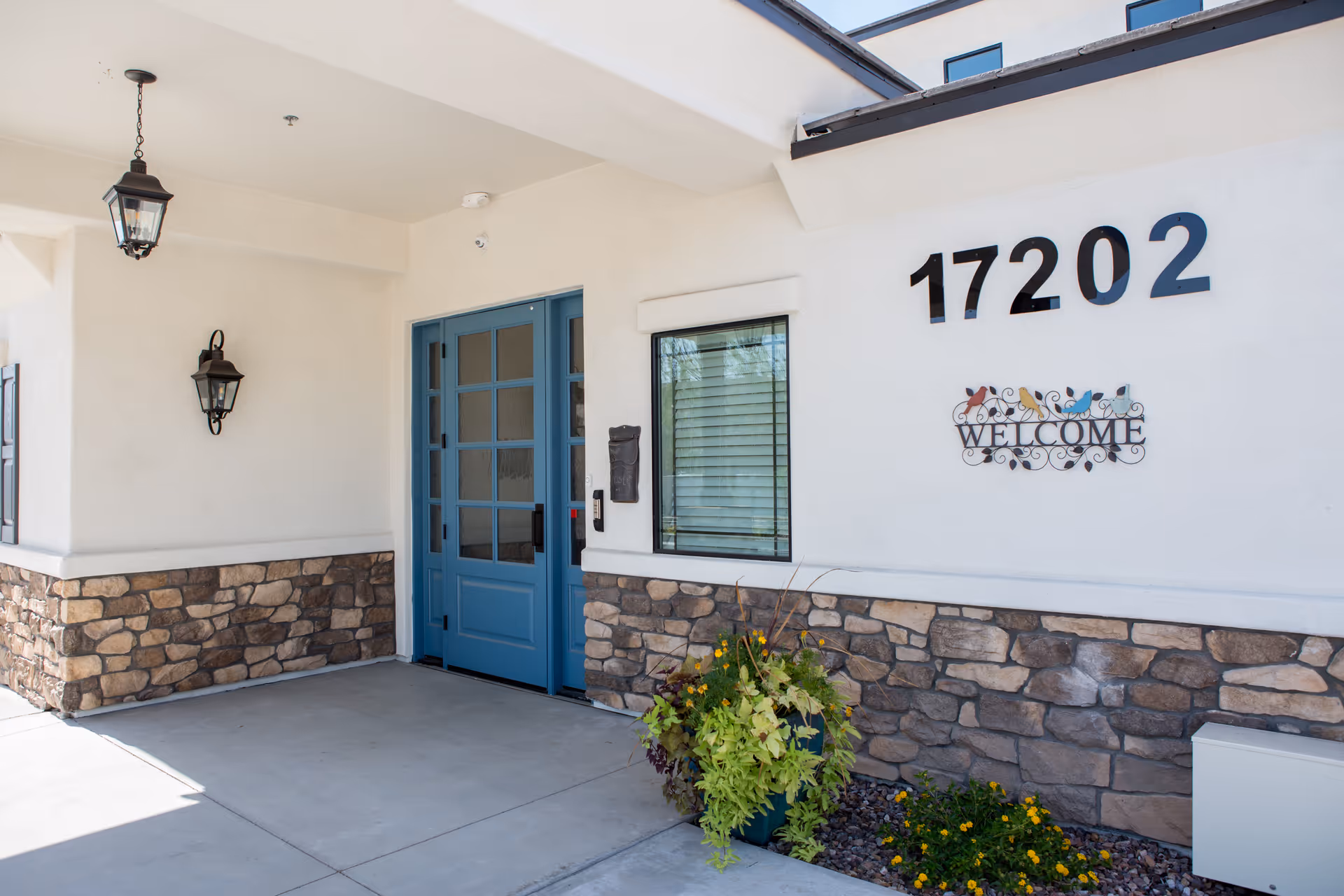 Entrance of a building with a blue double door, stone facade on the lower half of the wall, a window with blinds, and the number 17202 displayed above a decorative welcome sign with colorful birds. There are two black lantern-style light fixtures and a planter with green and yellow plants near the door.