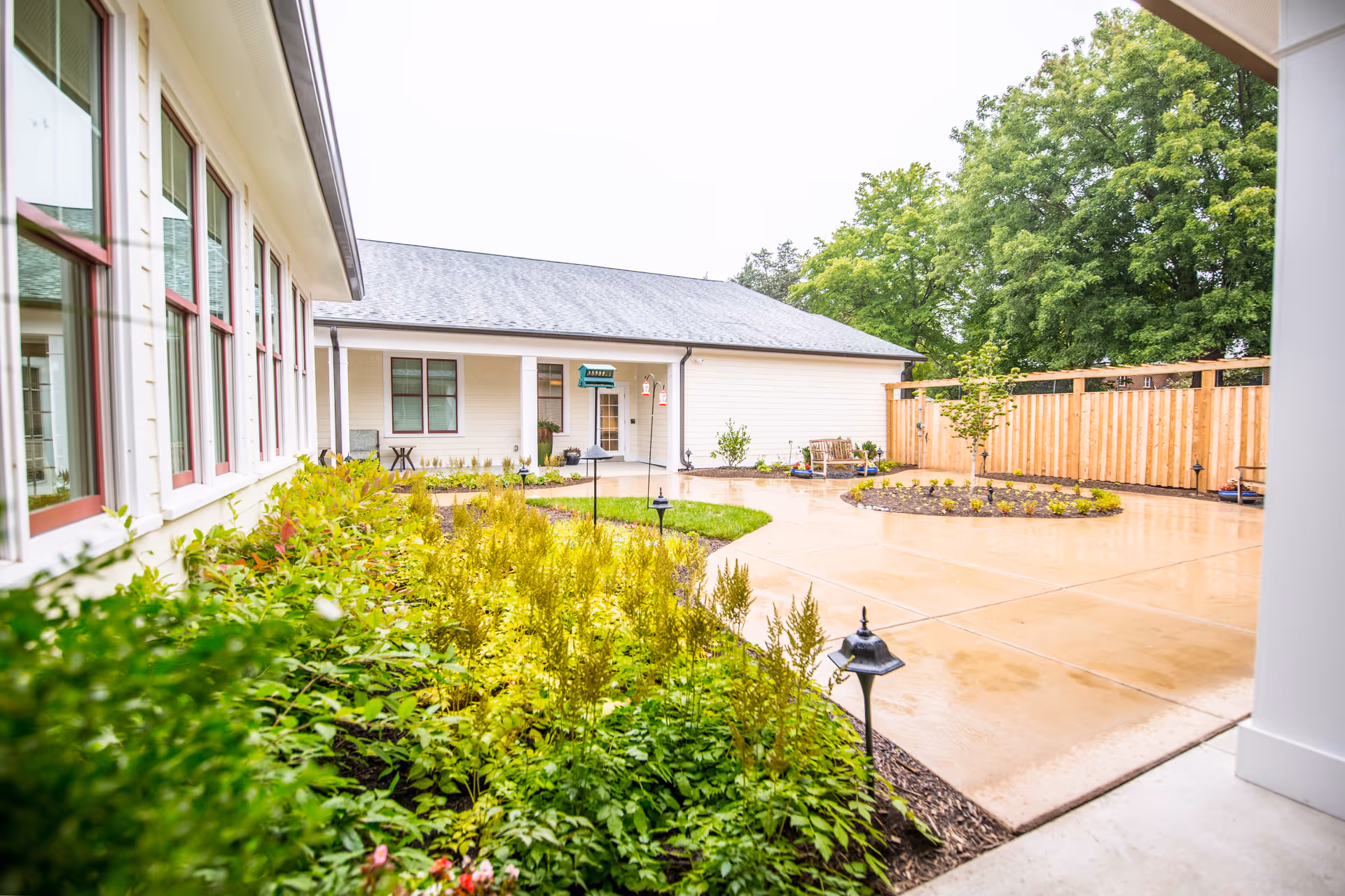 Outdoor courtyard area of Chantilly Heights Memory Care featuring a paved walkway, green plants and shrubs, a wooden fence, and a building with white siding and multiple windows.