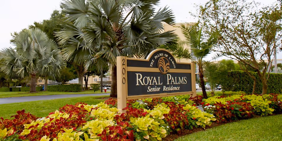 A landscaped garden area with colorful red and yellow flowers in front of a sign that reads 'Royal Palms Senior Residence' with palm trees and other greenery in the background.