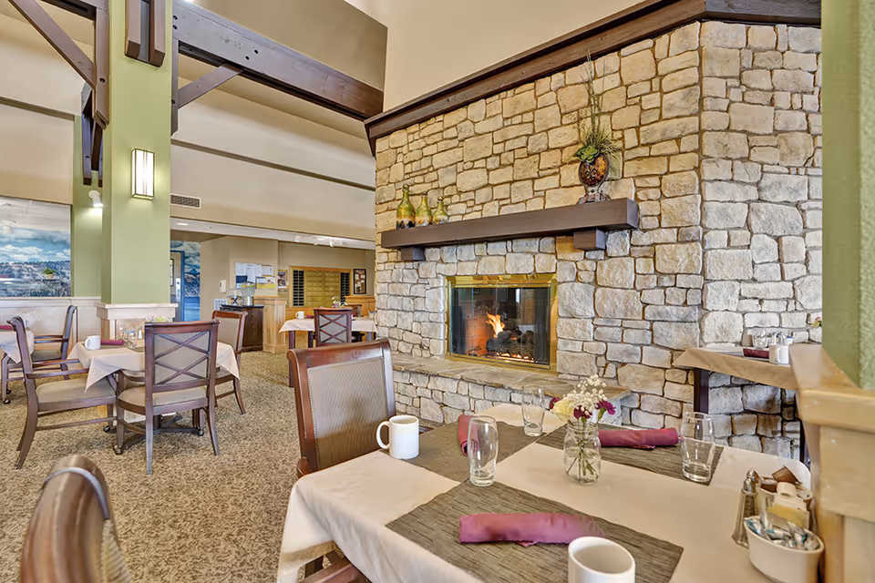 A cozy dining area in a senior living facility featuring a stone fireplace with a fire burning. Several tables are set with white tablecloths, purple napkins, glassware, coffee mugs, and small flower arrangements. The room has high ceilings with exposed wooden beams and soft lighting, creating a warm and inviting atmosphere.