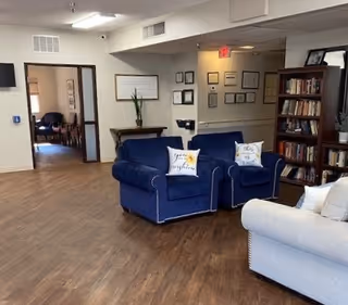 Interior view of a common area in a senior living facility with wooden flooring, blue and light gray upholstered armchairs with decorative pillows, a bookshelf filled with books, framed certificates on the wall, and an open doorway leading to another room with additional seating.