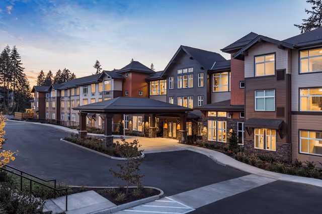 Exterior view of a large, multi-story senior living facility building at dusk with lights glowing from the windows. The building has a covered entrance with stone pillars and a driveway leading up to it. Trees and landscaping surround the area.