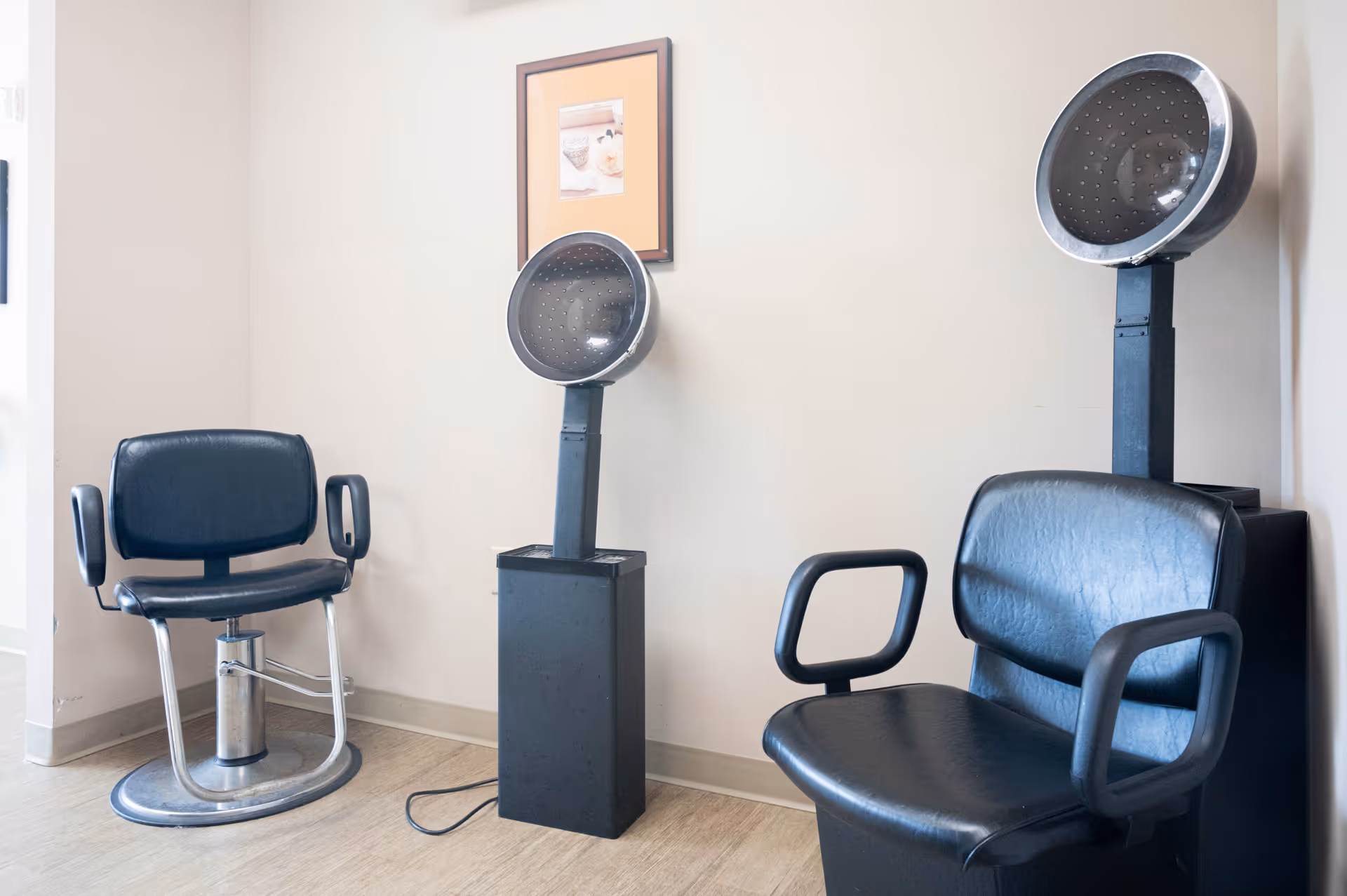 Interior view of a hair salon area with two black salon chairs and two vintage hair dryers mounted on stands against a beige wall with a framed picture.