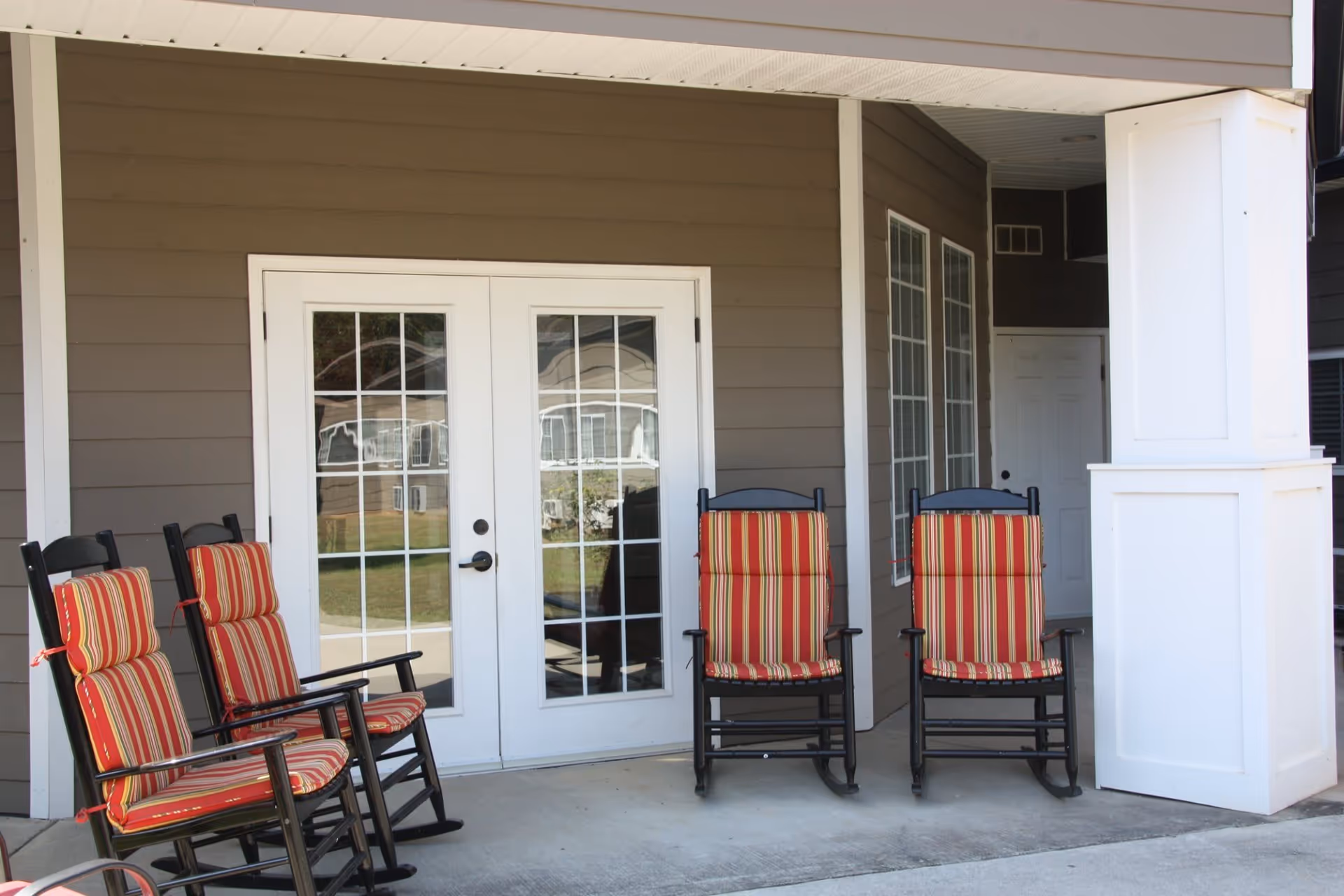 Outdoor patio area with four black rocking chairs featuring red and yellow striped cushions, positioned in front of a set of white double doors with glass panes, attached to a brown building with white trim.
