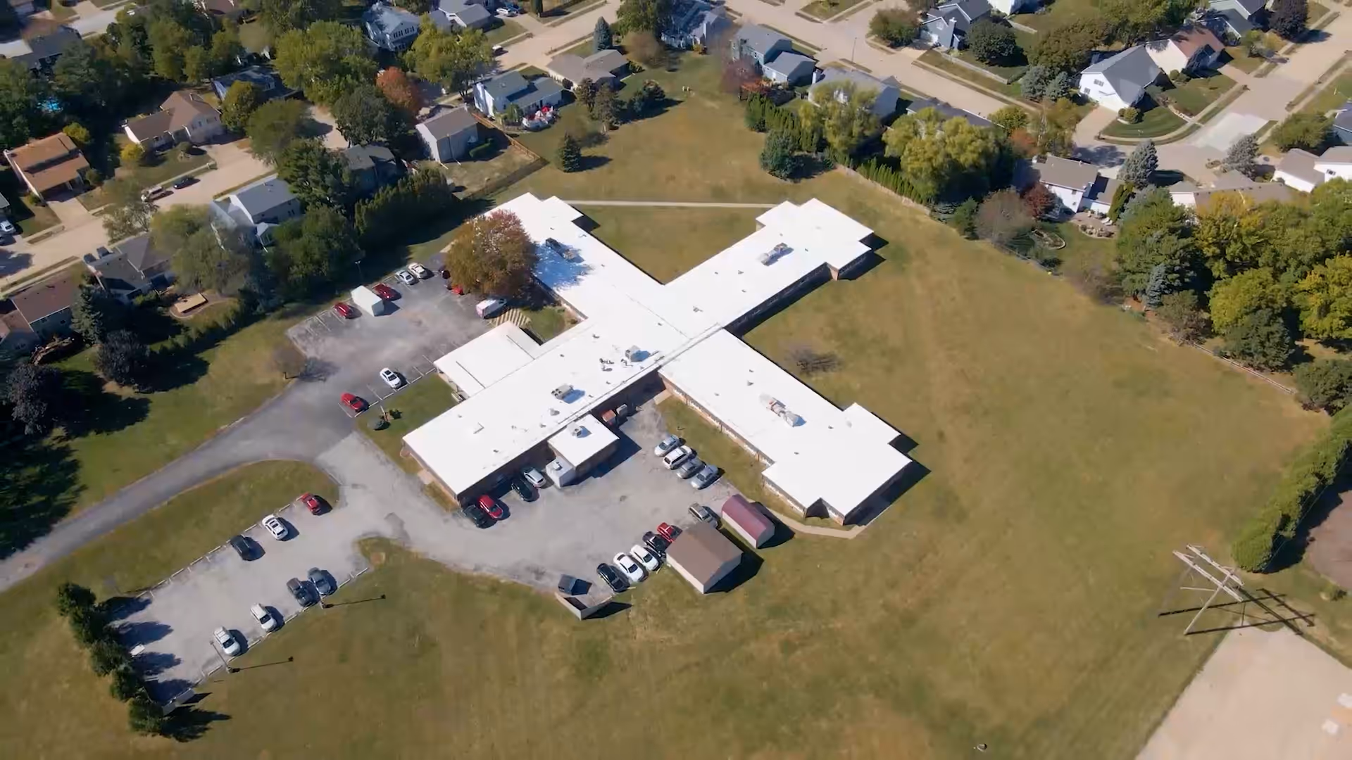 Aerial view of a cross-shaped single-story healthcare building with a parking lot, surrounding lawns, and nearby houses.