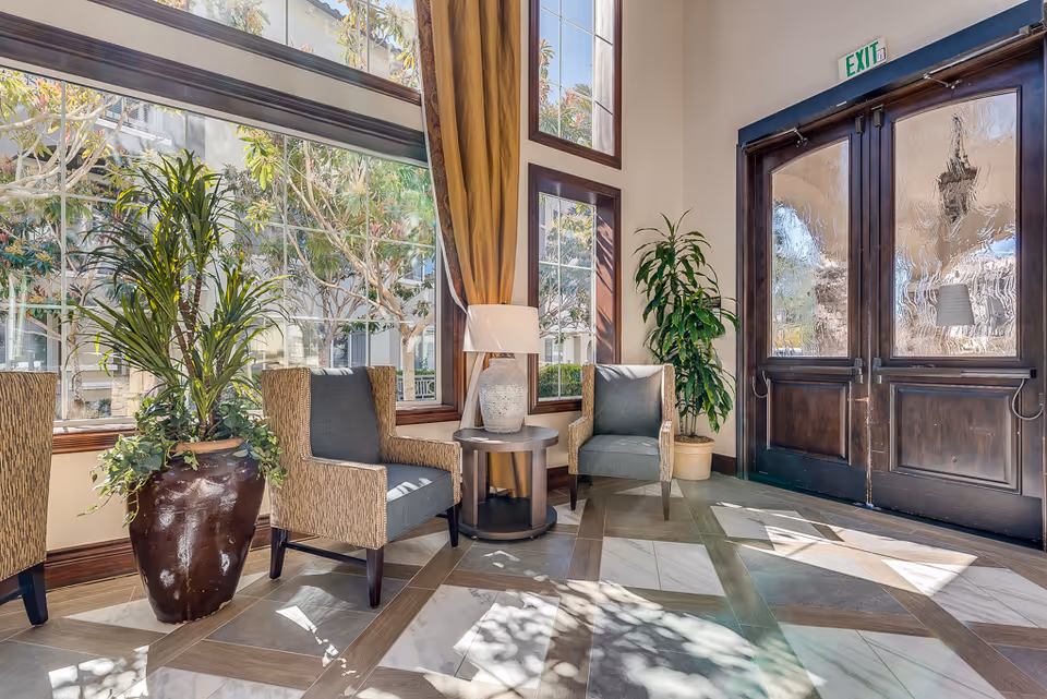 A bright and inviting seating area near large windows with natural light streaming in. The area features two cushioned armchairs with patterned upholstery, a round wooden side table with a decorative lamp, and two large potted plants. The floor has a geometric tile pattern, and there is a double wooden door with textured glass panels and an exit sign above it.
