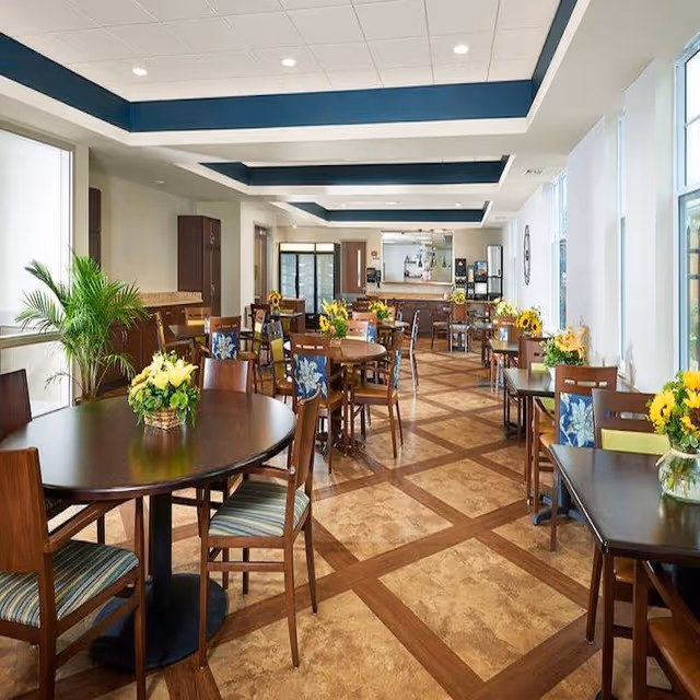 A bright and spacious dining room with multiple wooden tables and chairs arranged neatly. Each table has a floral centerpiece with yellow flowers. Large windows on the right side allow natural light to fill the room. The ceiling features recessed lighting and a blue accent design. There is a counter area at the far end of the room with additional seating and kitchen equipment visible behind it.