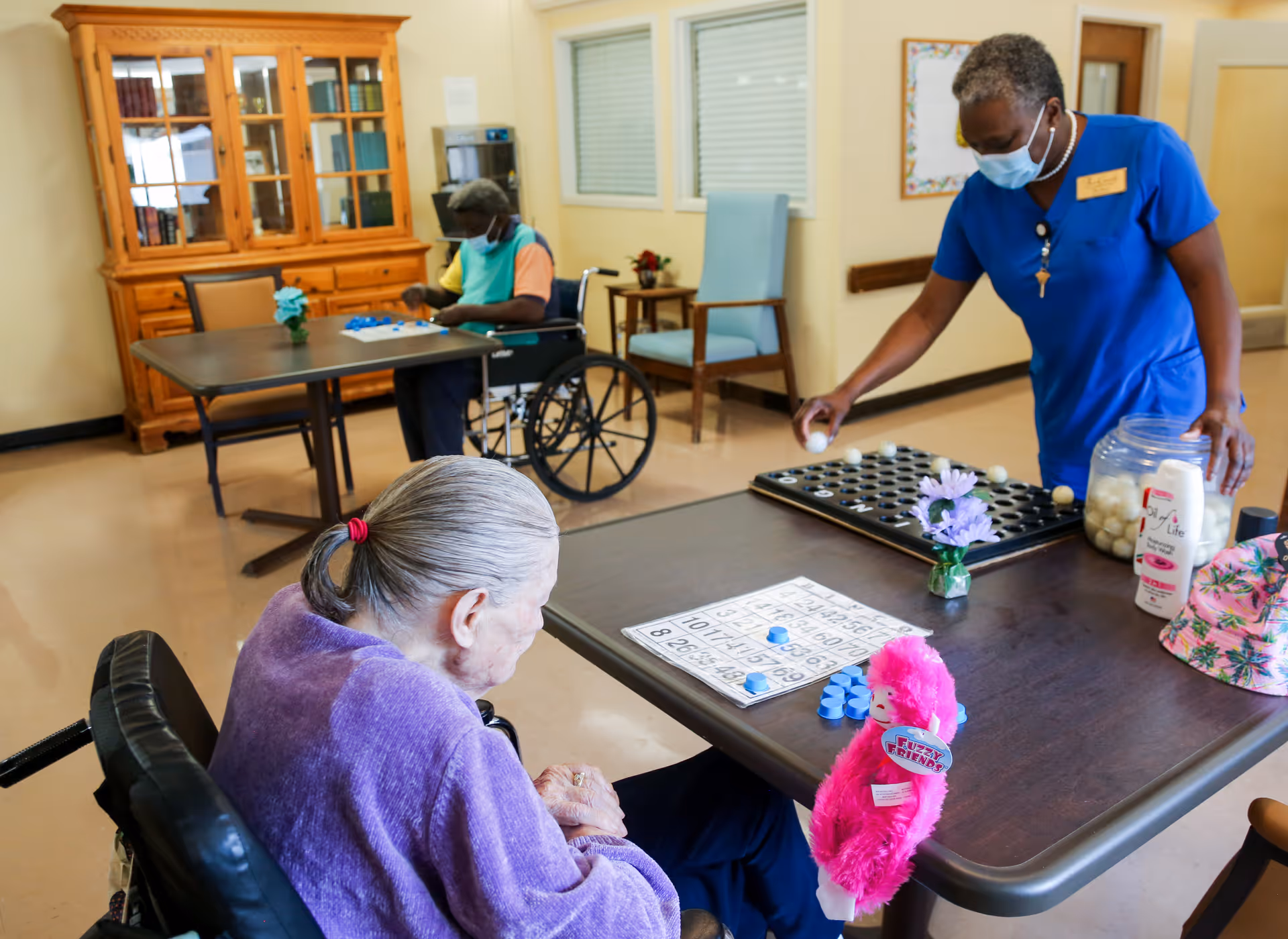 An elderly woman in a wheelchair playing bingo at a table with bingo cards and blue markers, while a staff member in blue scrubs and a face mask places bingo balls on a board. Another elderly person in a wheelchair is playing a game at a separate table in the background. The room has light-colored walls, a wooden cabinet with books, and chairs.