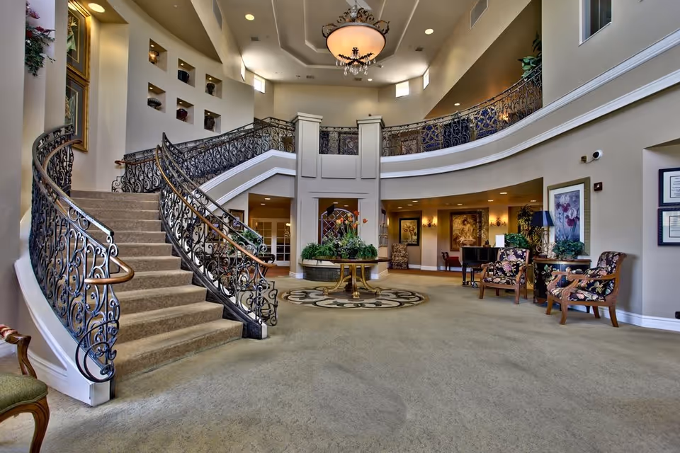 Spacious and elegant interior lobby area with a grand curved staircase featuring ornate wrought iron railings. The room has high ceilings with a large chandelier, decorative plants on a round table in the center, and several upholstered chairs with floral patterns arranged near framed artwork on the walls.