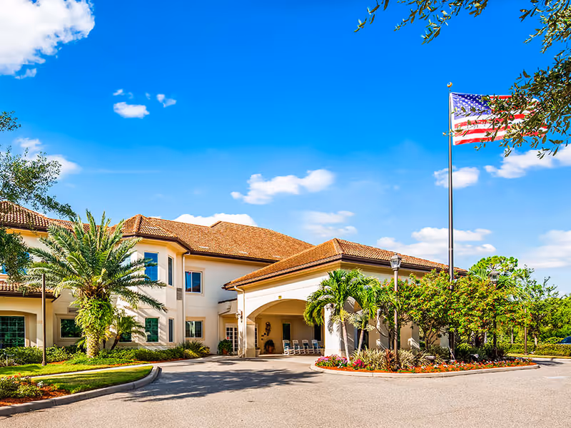 Front entrance of a two-story senior living building with a covered driveway, palm trees, landscaping, and an American flag flying on a tall flagpole.