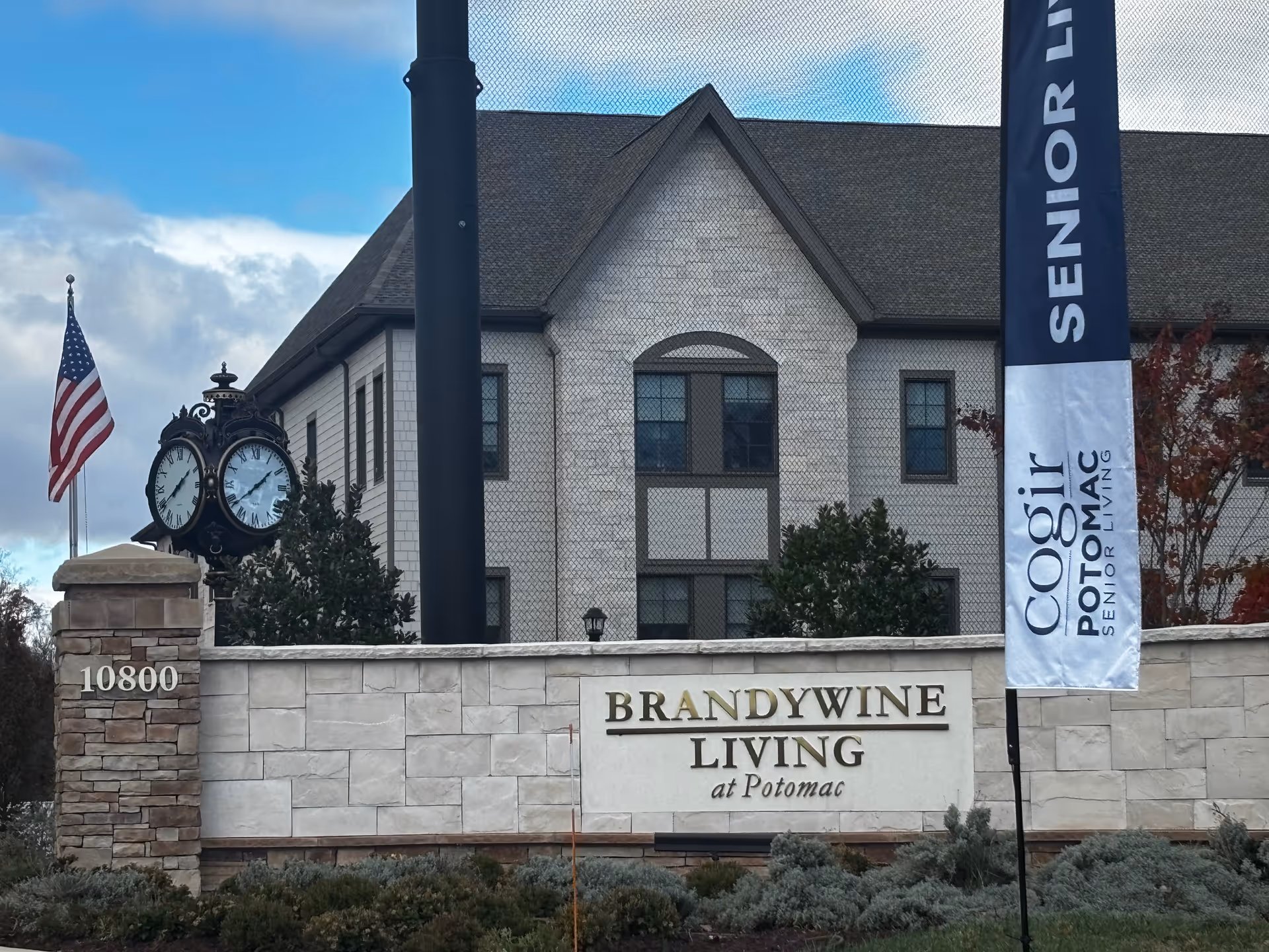 Stone entrance sign reading "Brandywine Living at Potomac" in front of the facility building with a decorative clock, American flag, and a vertical senior living banner.