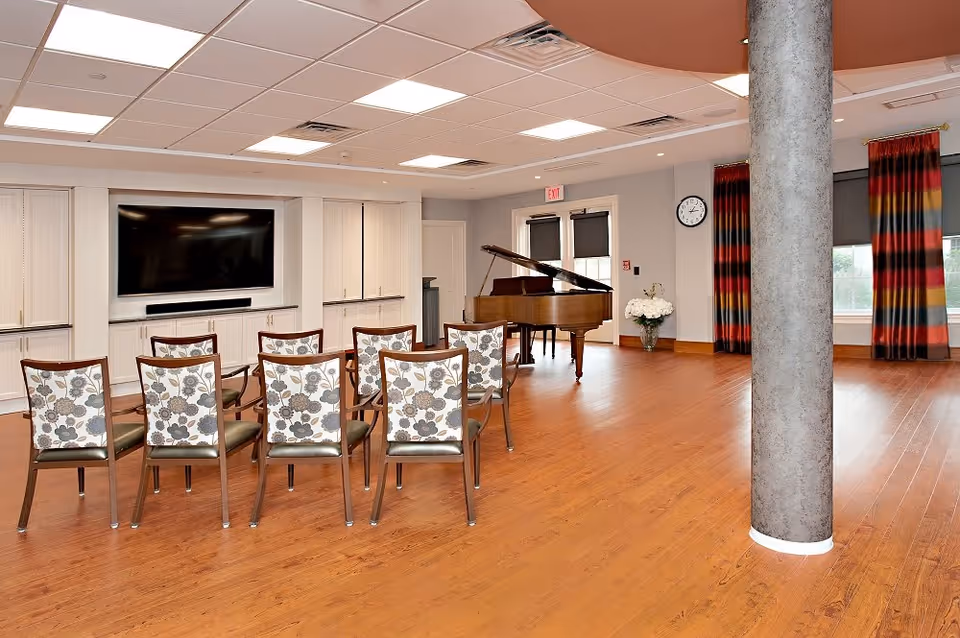 A spacious room with wooden flooring featuring a row of eight chairs with floral patterned backs facing a large flat-screen TV mounted on a white built-in cabinet. A grand piano is positioned near double doors with windows, and colorful striped curtains frame the windows on the right side. A round clock is mounted on the wall above a vase of white flowers, and a large cylindrical column stands in the foreground.