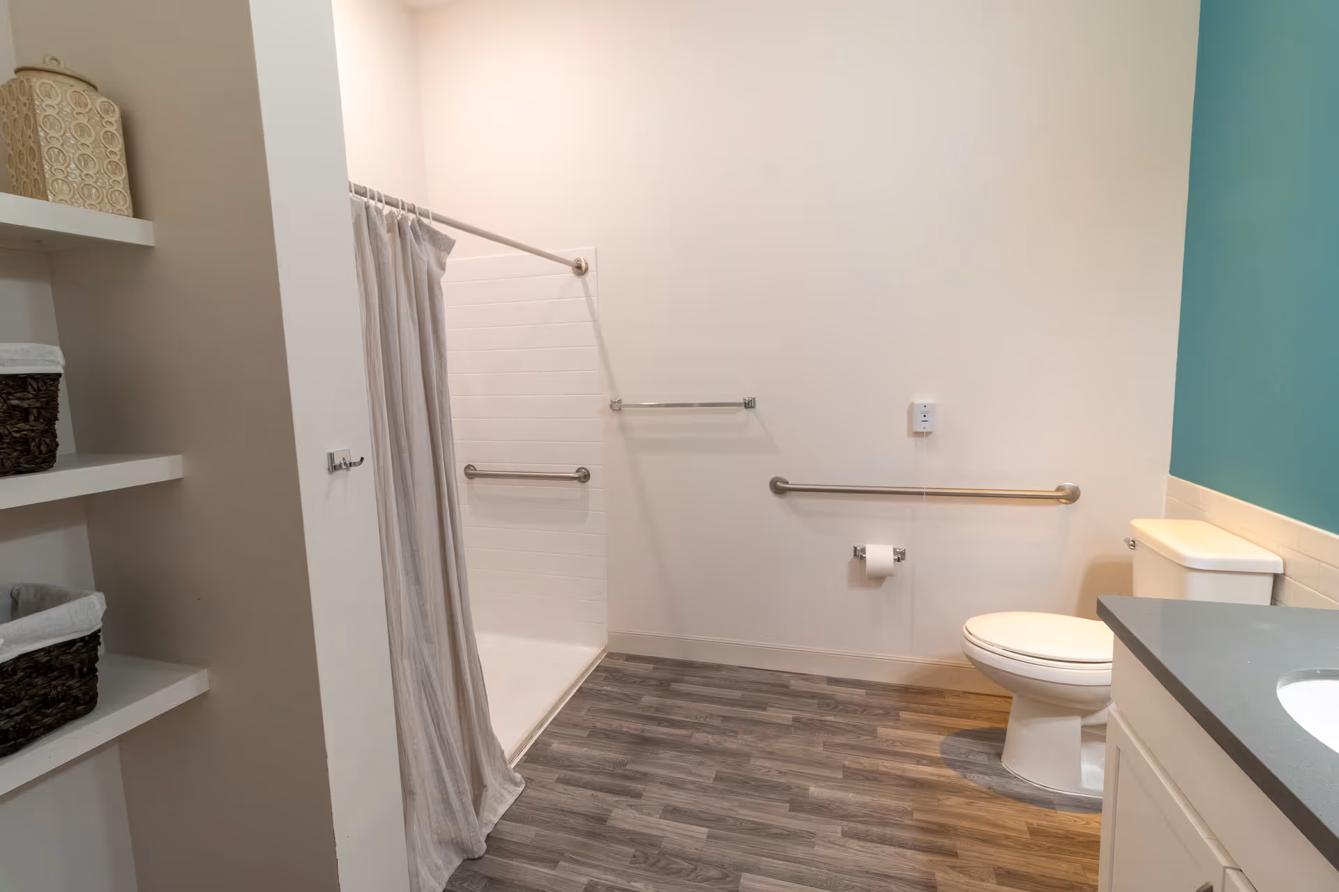 Accessible bathroom with a walk-in shower and curtain, toilet with grab bars, shelving, and a sink countertop.