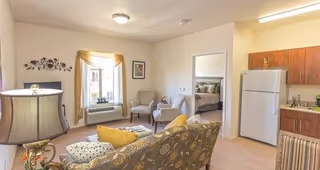 A cozy living area in a retirement facility featuring a patterned sofa with yellow and patterned cushions, two beige armchairs, a window with a yellow valance, a floor lamp, and a small kitchen area with wooden cabinets, a white refrigerator, and a countertop. A bedroom with a bed is visible through an open doorway.