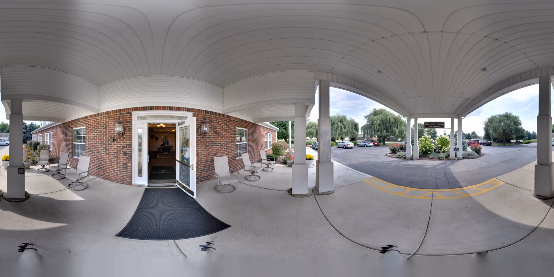Covered entrance area of a brick building with white pillars and ceiling. Several chairs are arranged along the wall near the entrance door, which is open. Outside the covered area, there is a parking lot with several cars and landscaped greenery including trees and bushes. A sign is visible near the parking lot entrance.
