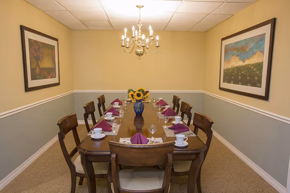 A formal dining room with a long wooden table set for eight people. Each place setting includes a white cup and saucer, a wine glass, silverware, and a folded purple napkin. A blue vase with sunflowers is centered on the table. The room has beige and light blue walls with two framed paintings and a chandelier hanging from the ceiling.