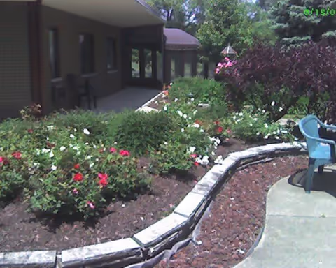 Outdoor garden area with a curved stone border, flower beds with red and white flowers, green bushes, and a blue plastic chair on a concrete patio. A building with a covered porch is visible in the background.
