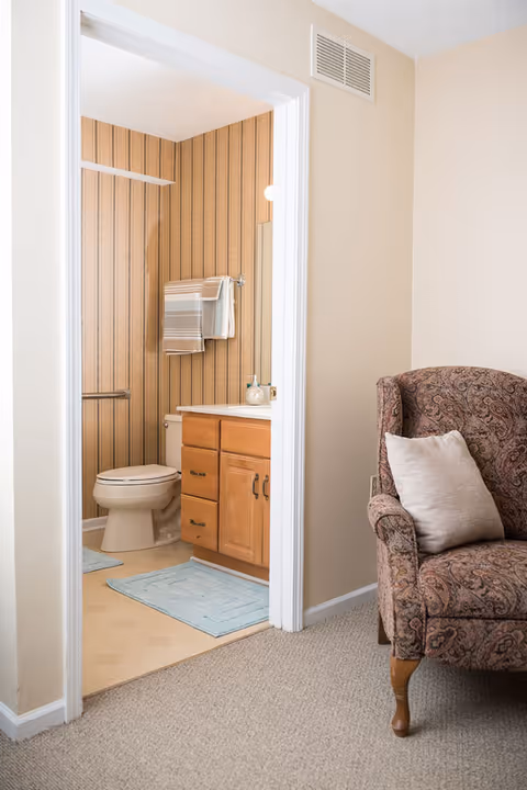 View of a bathroom with a toilet, wooden vanity with drawers, and a mirror. The bathroom walls have vertical striped wallpaper. There are two blue bath mats on the floor and two folded towels hanging on a towel rack above the toilet. To the right of the bathroom doorway, there is a patterned upholstered armchair with a light-colored pillow in a carpeted room.