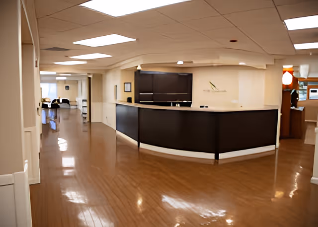 Interior view of a senior living facility's reception area with a curved dark wood front desk, light-colored walls, and a polished wooden floor. The hallway extends to the left with seating visible in the distance.