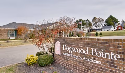 Exterior view of Dogwood Pointe assisted living facility with a brick sign in the foreground displaying the facility's name and a building with trees and a lawn in the background under a cloudy sky.