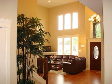 A bright living room with high ceilings and large windows letting in natural light. The room features brown leather sofas arranged around a wooden coffee table on a rug. There is a tall indoor plant near a staircase, a floor lamp, and a chandelier hanging near the entrance door with an oval glass design.