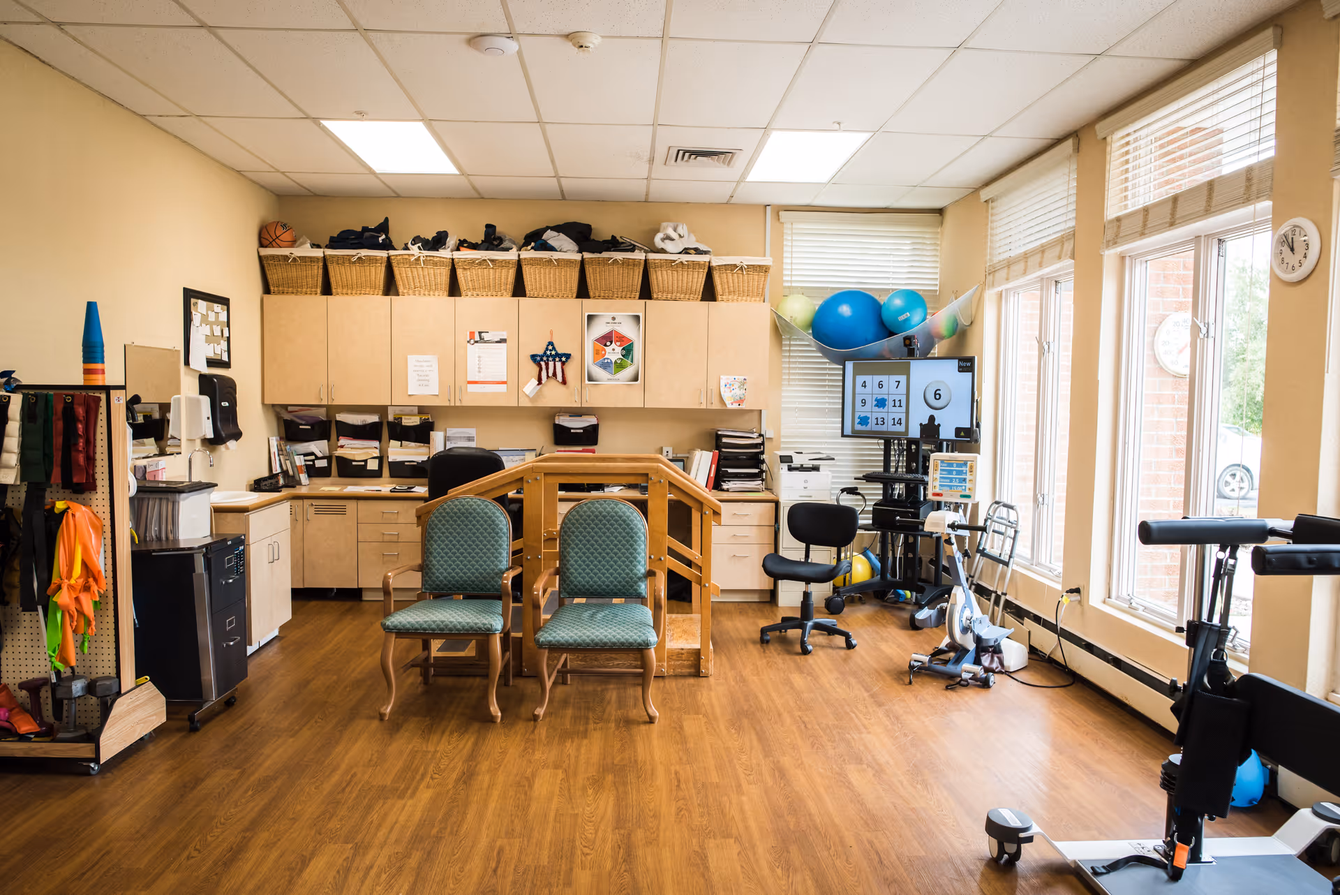A rehabilitation therapy room with wooden flooring, two green cushioned chairs, a wooden handrail structure, exercise equipment including a stationary bike and therapy balls, cabinets with baskets on top, and large windows letting in natural light.