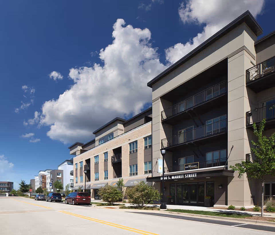 Exterior view of a modern multi-story building under a blue sky with scattered clouds. The building has balconies and large windows, with a sign that reads 'The Waters of Oak Creek' above the entrance. Several cars are parked along the street in front of the building.