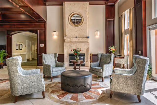 A cozy seating area with four gray upholstered armchairs arranged around a round black ottoman on a patterned tile floor. Behind the chairs is a large stone fireplace with a round mirror above it and two wall sconces on either side. The room features dark wood paneling and large windows letting in natural light. There are small tables with flower arrangements next to the chairs and in the background, a hallway and additional seating are visible.