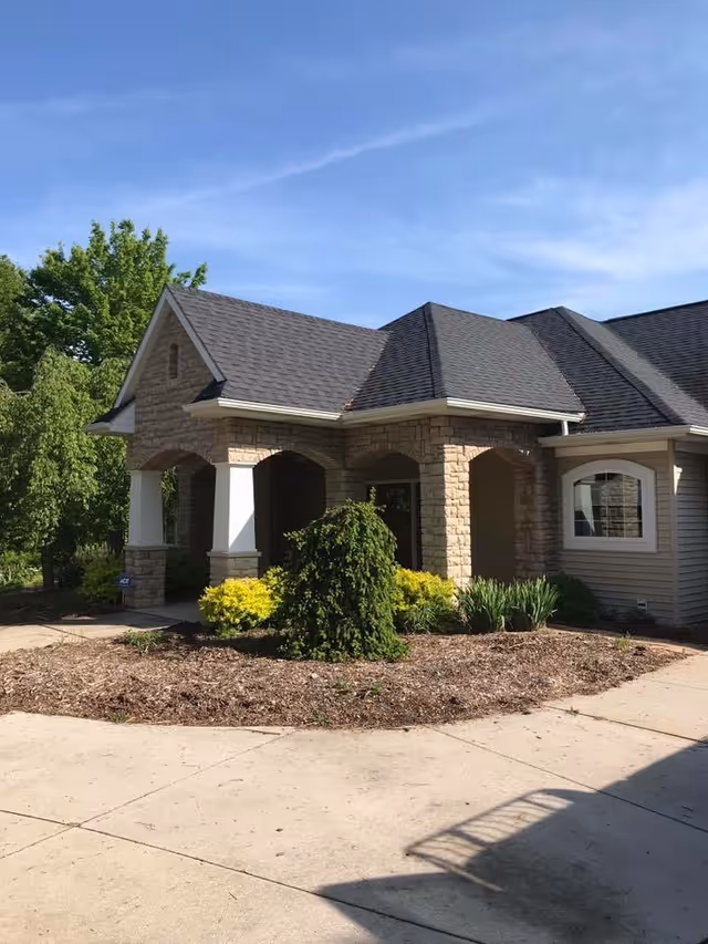 Front entrance of a one-story stone and siding building with arched porch, landscaping, and driveway under a blue sky.